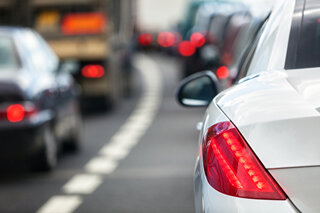 Das Bild zeigt einen Stau auf der Straße, mit einem weißen Auto im Vordergrund und verschwommenen Fahrzeugen im Hintergrund.