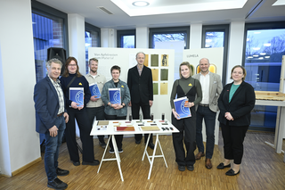 Das Bild zeigt eine Gruppe von Menschen, die stolz ihre Zertifikate halten. Im Hintergrund sind verschiedene Materialien und Muster ausgestellt.