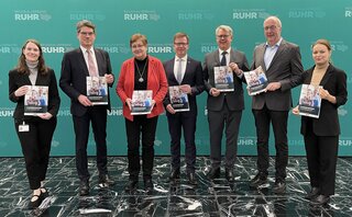 Die Gruppe von acht Personen steht vor einem grünen Hintergrund und hält Broschüren in den Händen. Sie wirken freundlich und professionell.