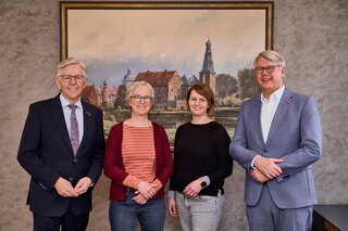 Das Bild zeigt vier Personen in einem Innenraum vor einem Kunstwerk. Sie lächeln und stehen freundschaftlich zusammen.