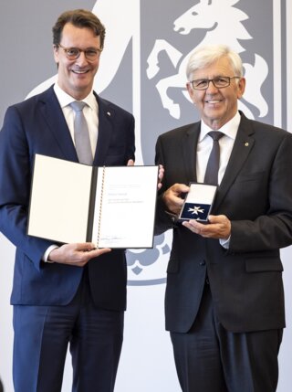 Die beiden Männer halten ein Dokument und eine Medaille. Sie lächeln in einer formellen Umgebung mit einem Pferdelogo im Hintergrund.
