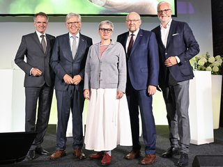 Fünf Personen stehen auf einer Bühne, lächeln und posieren für die Kamera. Sie tragen formelle Kleidung und wirken freundlich.
