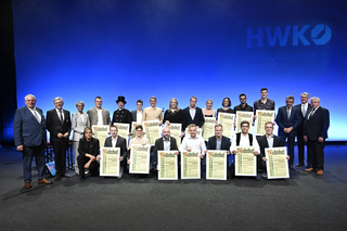 Eine Gruppe von jungen Handwerkern steht stolz mit Urkunden auf der Bühne. Im Hintergrund ist die HWK-Logo und eine festliche Atmosphäre zu sehen.